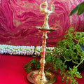 Traditional brass kuthu vilakku (oil lamp) with a peacock design, surrounded by plants and vibrant decor on a red surface.