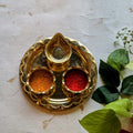 Intricately designed brass kumkum plate with compartments for kumkum and turmeric, accompanied by a central holder, displayed with floral accents.