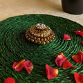 Round kumkum box in gold with ornate design and crystal accents, placed on a green woven mat with scattered rose petals.