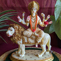 Handcrafted Durga idol in ivory and gold finish, seated on a lion with eight arms holding divine weapons, displayed on a green mat with a red backdrop and lush foliage.