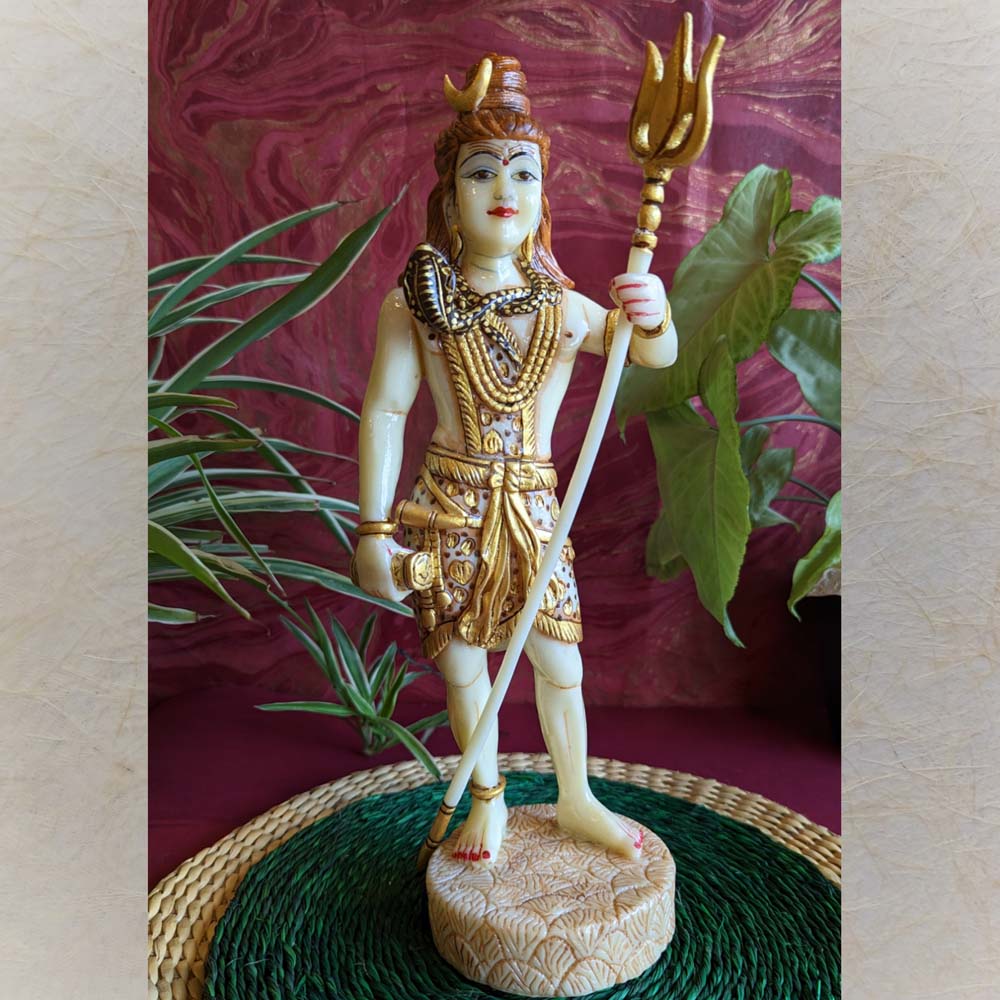Handcrafted Standing Shiva idol in ivory and gold finish, holding a trident, with intricate detailing, displayed on a green mat with a red background and foliage.