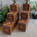 Three-step wooden Vilakku Padi with white kolam hand-painted designs, crafted from teak wood, displayed with lamps and plants.