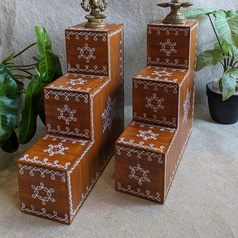 Three-step wooden Vilakku Padi with white kolam hand-painted designs, crafted from teak wood, displayed with lamps and plants.