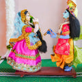 Handcrafted dolls of Radha and Krishna in a playful pose, with Radha offering a betel leaf to Krishna, adorned in vibrant traditional attire with intricate embellishments.