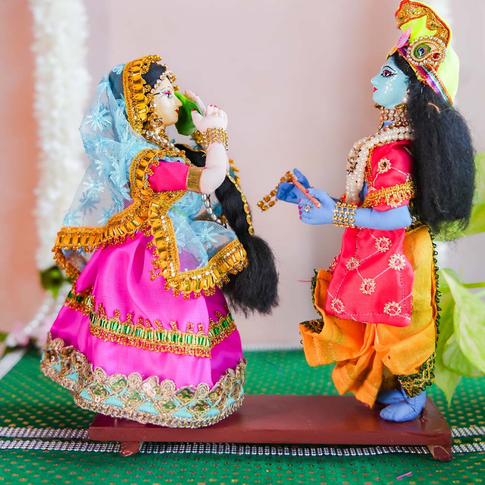 Handcrafted dolls of Radha and Krishna in a playful pose, with Radha offering a betel leaf to Krishna, adorned in vibrant traditional attire with intricate embellishments.