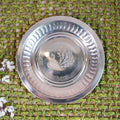 Stainless steel plate with an etched peacock design at the center, placed on a woven green background with floral accents.