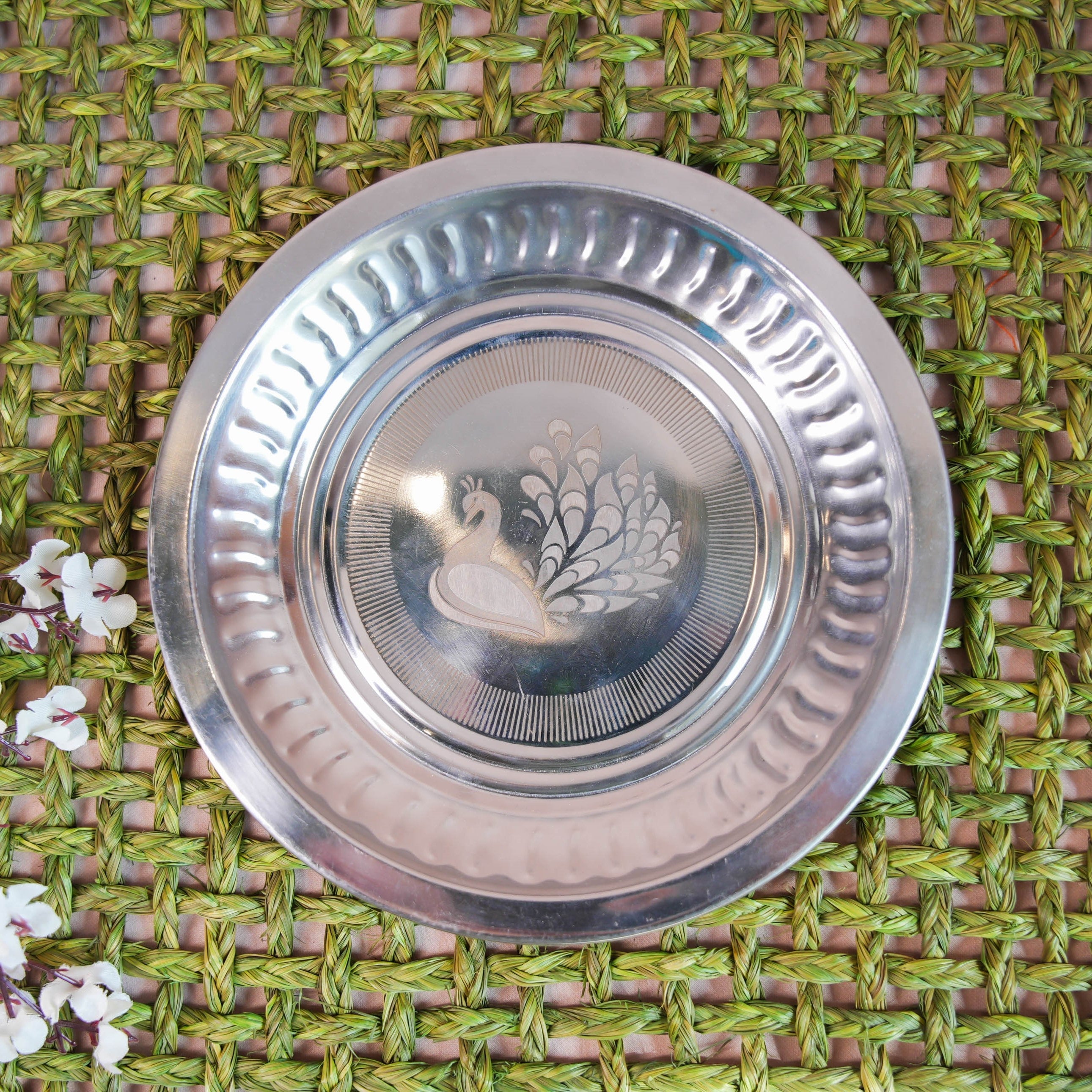 Stainless steel plate with an etched peacock design at the center, placed on a woven green background with floral accents.