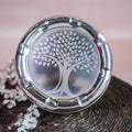 Stainless steel laddu box with a laser-etched tree design on the lid, placed on a woven mat surrounded by floral decor.