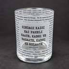 A clear glass tumbler with printed black text, featuring the Hindi phrase 'ZINDAGI KAISI HAI PEHeli HAAYE, KABHI YE HASAYE, KABHI YE RULAAYE' and the name 'Anand (1970)', set against a dark gray background.