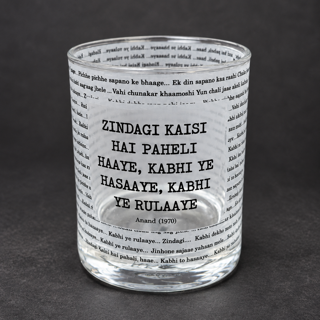 A clear glass tumbler with printed black text, featuring the Hindi phrase 'ZINDAGI KAISI HAI PEHeli HAAYE, KABHI YE HASAYE, KABHI YE RULAAYE' and the name 'Anand (1970)', set against a dark gray background.