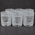 Six clear glass shot glasses arranged in two rows on a dark gray surface. Each glass is printed with black text in a newspaper-style pattern, featuring Punjabi phrases such as 'Tum Tuna Jo' and 'Jeena Yaha'.