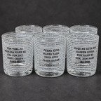 Six clear glass shot glasses arranged in two rows on a dark gray surface. Each glass is printed with black text in a newspaper-style pattern, featuring Punjabi phrases such as 'Tum Tuna Jo' and 'Jeena Yaha'.