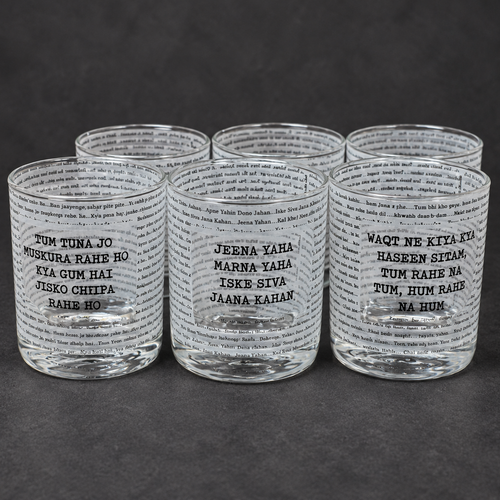 Six clear glass shot glasses arranged in two rows on a dark gray surface. Each glass is printed with black text in a newspaper-style pattern, featuring Punjabi phrases such as 'Tum Tuna Jo' and 'Jeena Yaha'.